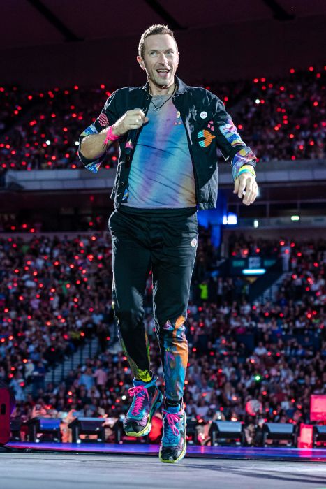 The singer delivers an energetic performance on stage, sporting a vibrant shirt and jacket, surrounded by a massive crowd and dazzling lights.