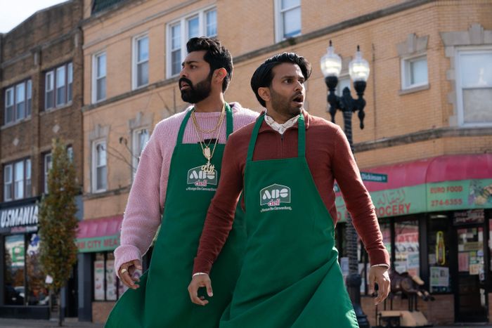 Two actors, dressed in green aprons, stand on a bustling street looking astonished, captured in a scene from a TV show or movie.