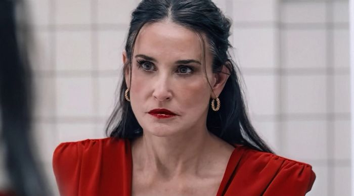 Demi Moore, with her long dark hair and bold gold hoop earrings, exudes a serious expression as she dons a chic V-neck top, gazing into a mirror in a tiled room. This scene is from her role in 