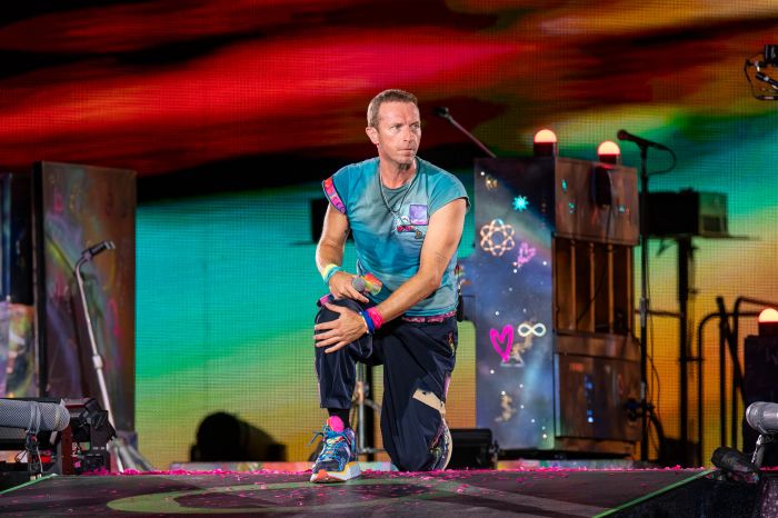 A performer kneels on stage during a lively concert, wearing casual, artistic clothing. Bright visuals and musical instruments can be seen in the background