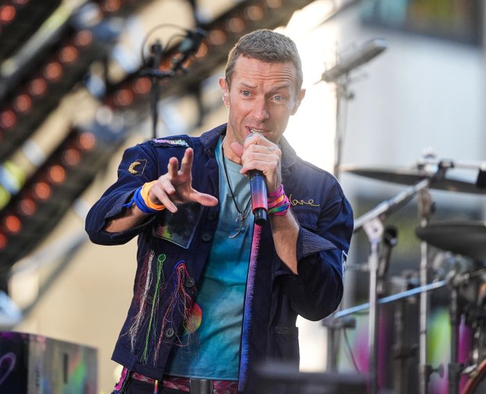 A performer on stage sings passionately while wearing a vibrant, multicolored jacket and shirt, energetically gesturing towards the crowd. Musical instruments are visible in the background