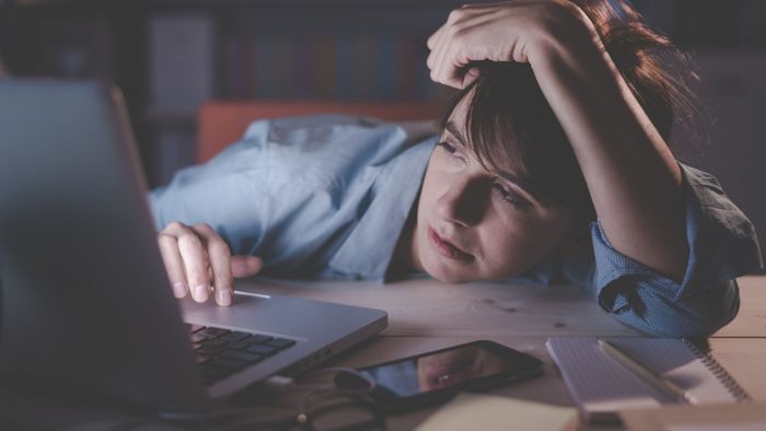 A photo of a tired woman resting her head on a desk while working on a laptop