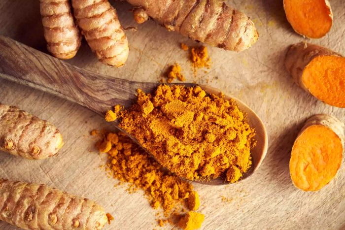 A spoonful of curcuma powder alongside fresh, whole, and sliced turmeric root, viewed from above