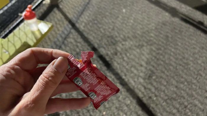 My hand grips an open ketchup packet as I stand on a track at night, with a water bottle visible on a bench in the background.