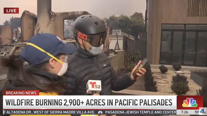 Two journalists, equipped with helmets and masks, stand in front of a building severely damaged by fire, providing live coverage of a wildfire in Pacific Palisades. News banners flash real-time updates.