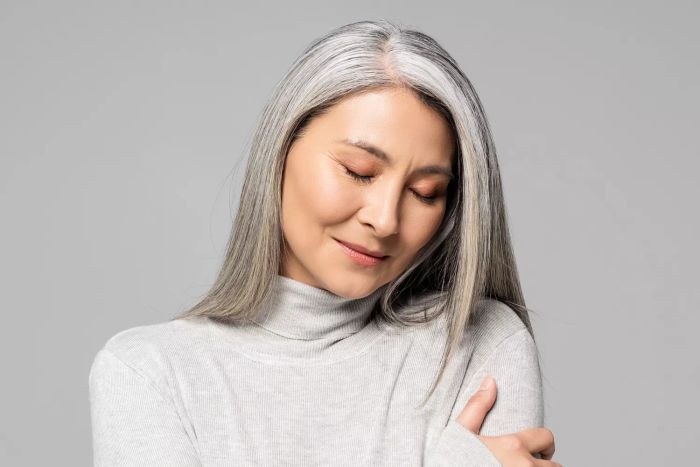 A serene Asian woman in a turtleneck, with grey hair and eyes gently closed, set against a grey backdrop.