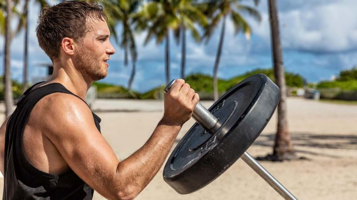A person is gripping a loaded barbell at shoulder height, with the opposite end angled downward and resting on a ground support or attachment, out of sight.