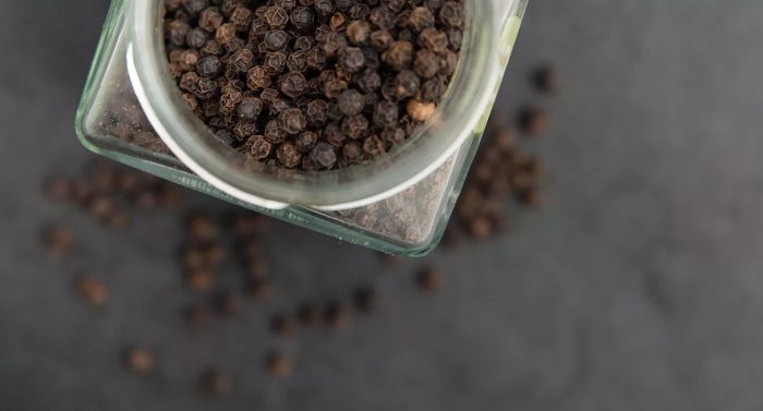 Black pepper (Piper nigrum). A glass jar filled with black peppercorns against a greyish backdrop.