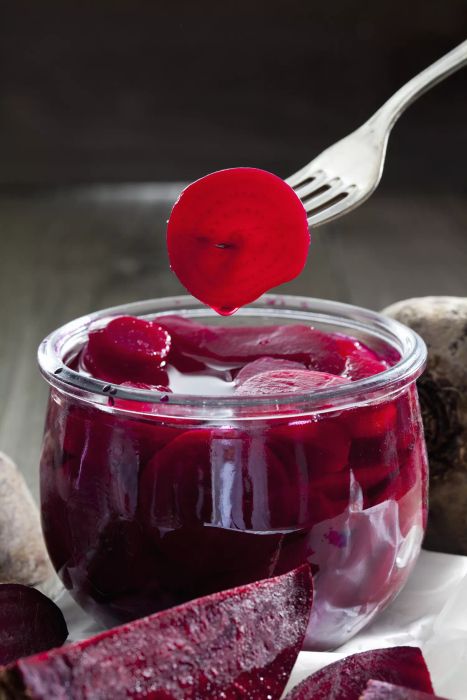 A small glass jar filled with slices of pickled beets, accompanied by a fork positioned above the jar, ready to skewer one of the slices.