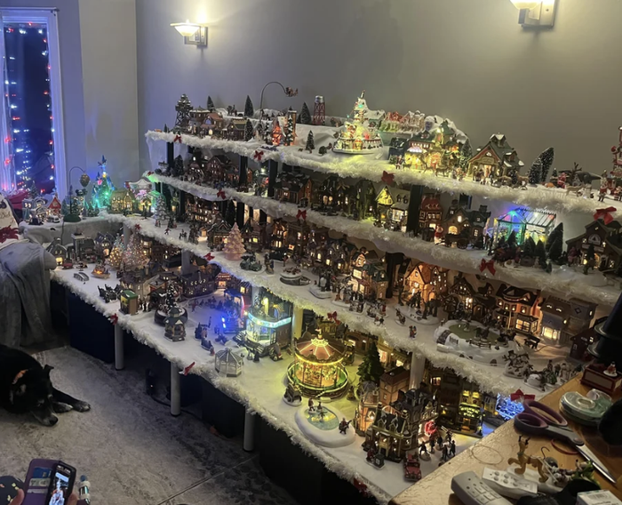 A holiday-themed miniature village display featuring lights, various buildings, snowy rooftops, and a central carousel.