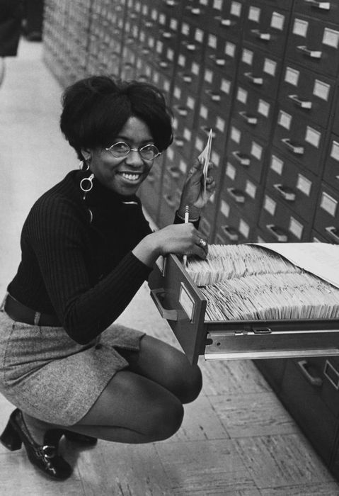 A woman wearing glasses crouches beside an open file drawer in a large filing cabinet system, holding cards in her hands, dressed in a turtleneck and skirt, smiling at the camera