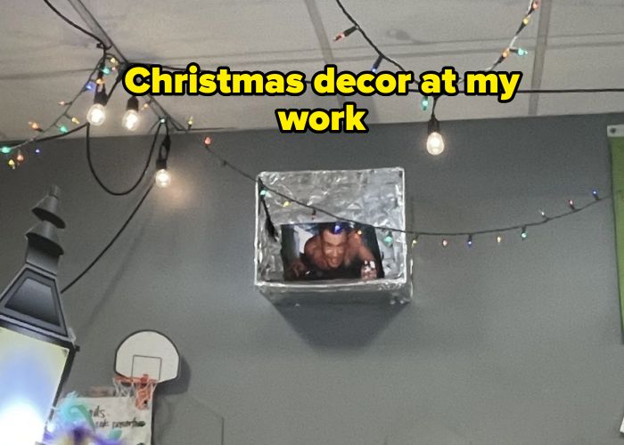 A man in a small metallic box seems to be emerging from the ceiling, playfully gesturing while surrounded by string lights, with part of a basketball hoop visible