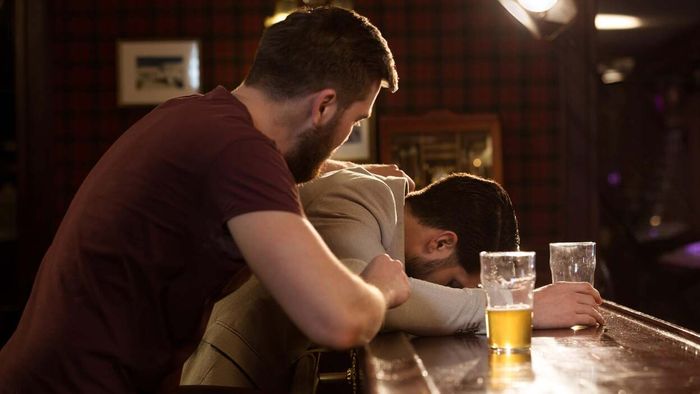 A worried individual checking on a friend who has passed out at the bar with an empty beer glass beside them