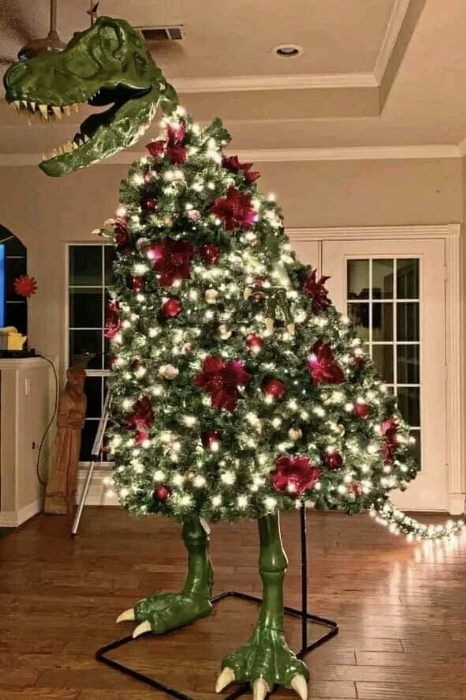 A dinosaur-shaped Christmas tree, complete with lights and red decorations, displaying a large open mouth and standing on green T-rex legs in a living room