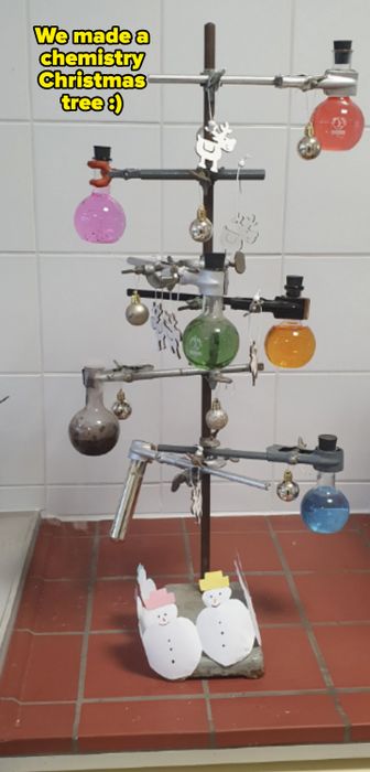 A chemistry-themed Christmas tree adorned with lab equipment, ornaments, and paper snowmen, placed against a tiled wall
