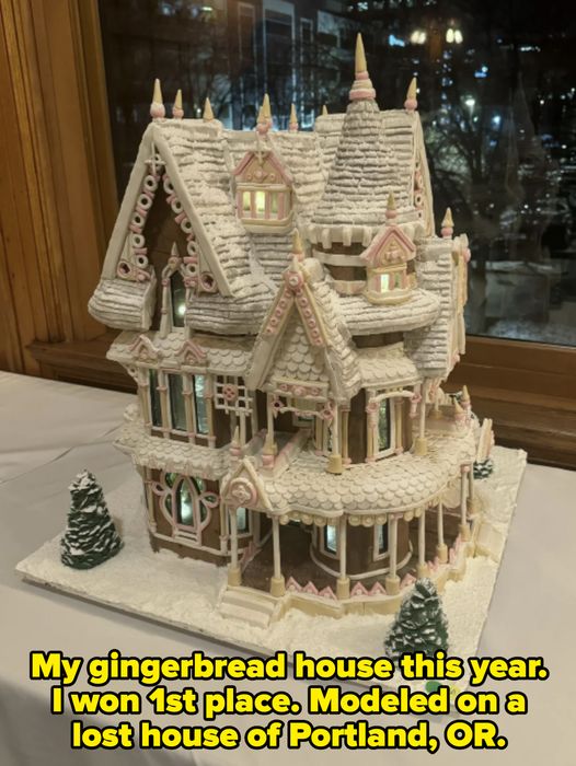 An intricately designed gingerbread house with turrets, a wraparound porch, and decorated with icing and candy, set on a snowy base with two small trees.