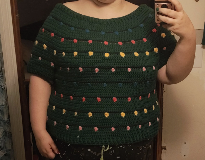 A person wearing a crocheted sweater adorned with colorful yarn dots, taking a mirror selfie.