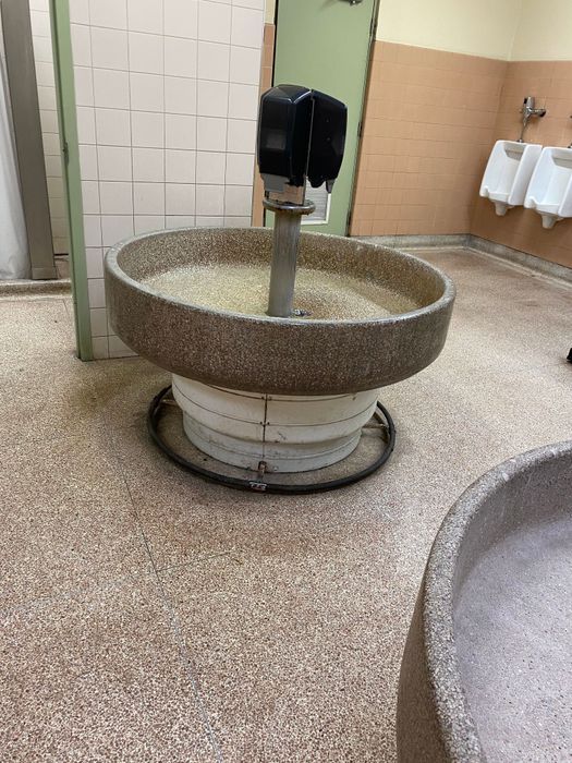 A round stone-wash bathroom sink with a touchless soap dispenser in a public restroom. Urinals and tiled walls are visible in the background.