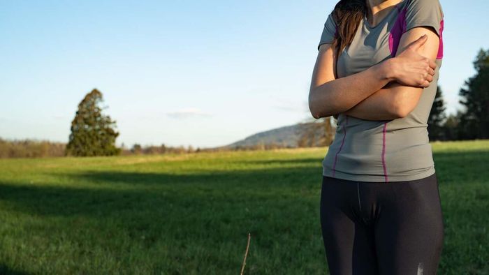 A woman dressed in workout attire wraps her arms around herself for warmth, standing outdoors on a bright sunny day