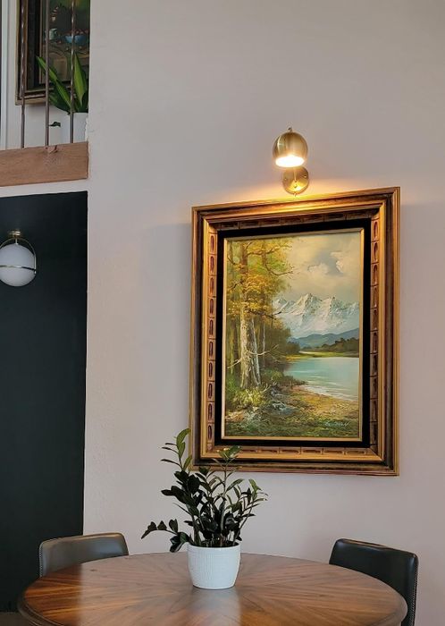 A framed landscape painting with trees, mountains, and a lake hangs above a round table with a potted plant below.