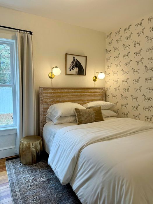 A bedroom featuring a wooden headboard, horse-themed wallpaper, and a neatly made bed with crisp white linens. Two wall sconces frame a horse portrait above the bed.