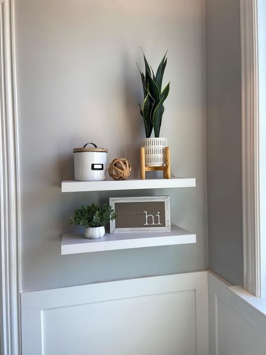 Two pristine white floating shelves, adorned with decorative items: a plant in a woven pot, a white canister, a ball of twine, a small plant, and a decorative sign with text.
