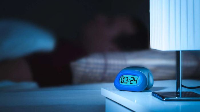 A close-up of a digital clock at night, with a person sleeping in the background.