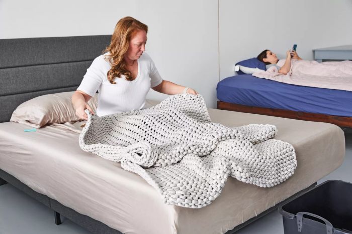 A person sitting on a bed covered with the Bearaby Cotton Weighted Blanket