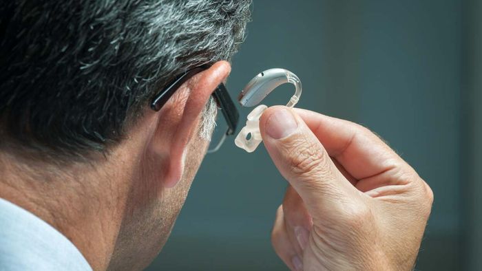 A senior man, seen from behind, shoulders up, taking off a hearing aid from his ear