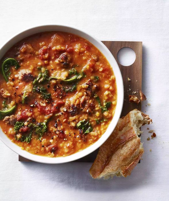 A hearty bowl of Lentil, Kale, and Sausage Soup served in a white dish atop a wooden board, accompanied by a slice of baguette.