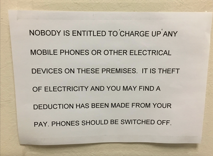 A sign reads: 'No one is permitted to 'charge' their mobile phones or any other electronic devices on these premises. Doing so will be considered theft of electricity, and you may notice a deduction from your pay. Please ensure your phones are switched off.'