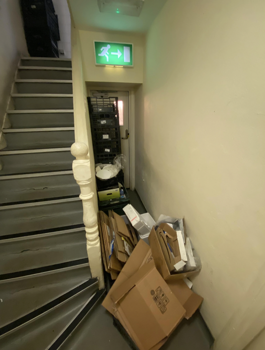 A fire escape door is obstructed by several cardboard boxes, baskets, and other items, blocking the staircase in a chaotic hallway