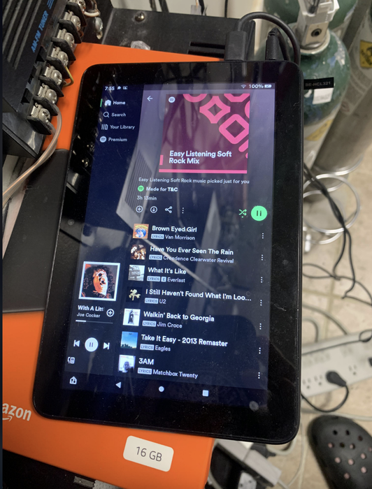 A tablet displays a Spotify playlist titled 'Easy Listening Soft Rock Mix,' surrounded by a messy setup with wires and an orange Amazon package