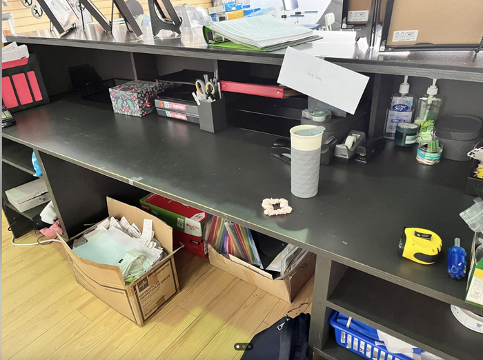 A messy office desk filled with papers, office tools, and personal items like a coffee mug, a tape measure, and a cardboard box full of documents on the floor