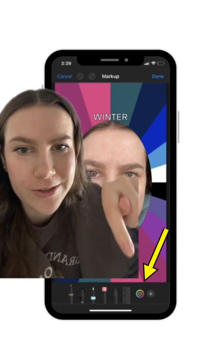 A woman points at her phone screen showing an app with the word 'WINTER' and a color wheel. The phone screen reflects her face.