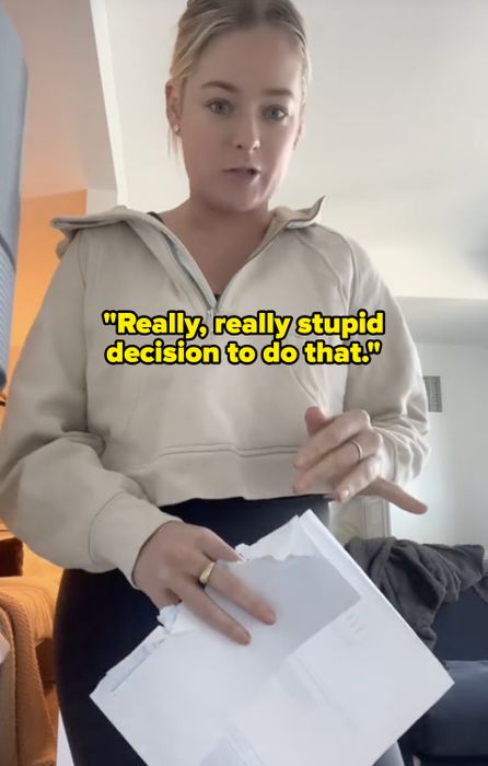 A woman standing indoors, dressed in a casual hoodie, holding and examining papers