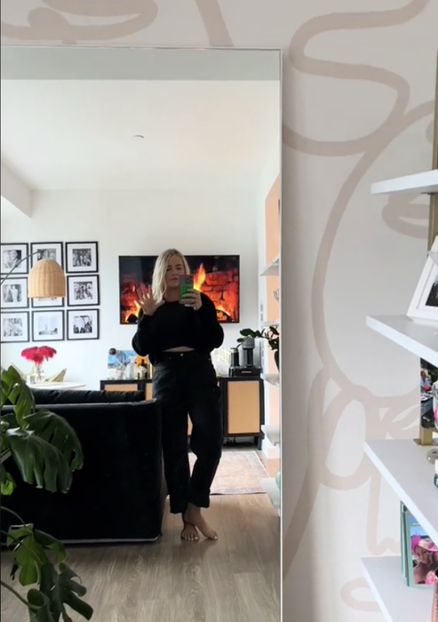A woman capturing a mirror selfie in a casual black outfit, with a living room visible in the background