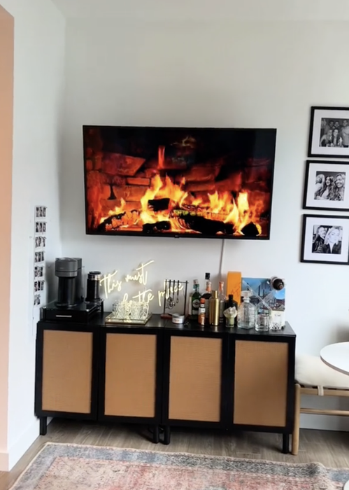 A TV showing a fireplace video, positioned above a cabinet stocked with bottles, a coffee machine, and framed photos on the wall