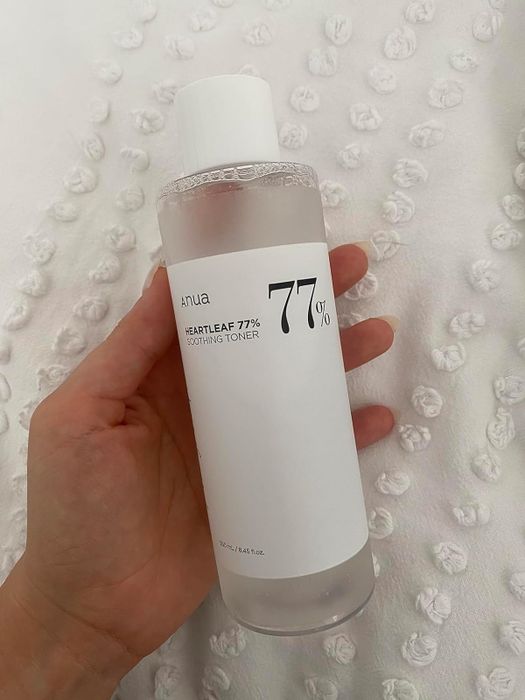 A reviewer holding the Anua Heartleaf 77% Soothing Toner against a textured backdrop