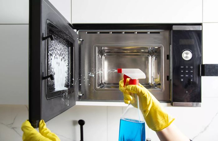 A woman using an anti-grease spray to clean the kitchen, focusing on wiping away grease and grime from the microwave. The modern anti-static cleaner helps keep surfaces spotless.