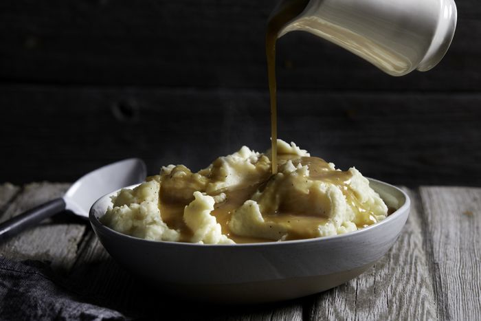 A jug of gravy being poured over a bowl of hot mashed potatoes.