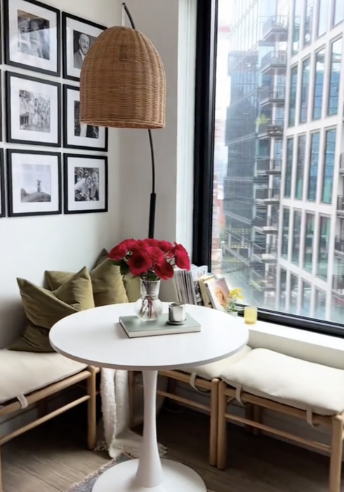 A snug corner with a small round table, two benches, a floor lamp, and wall frames next to a window offering a view of city buildings