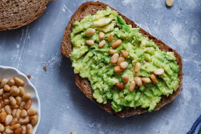 A top view of a rye bread sandwich topped with creamy avocado and roasted pine nuts, set against a textured blue background. A healthy eating concept.