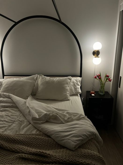 A softly lit bedroom featuring an unmade bed, a nightstand with a lamp, and a vase filled with tulips