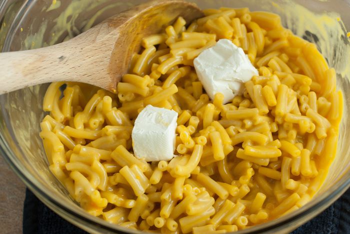 Boxed mac 'n' cheese being stirred with a wooden spoon in a bowl, with chunks of cream cheese visible.
