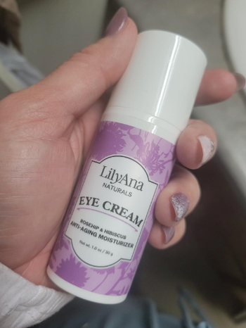 A person holding a LilyAna Naturals Eye Cream bottle, labeled as an anti-aging moisturizer with rosehip and hibiscus