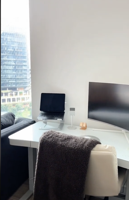 A home office setup with a laptop on a white desk, a monitor on the side, and a view of the city through the window