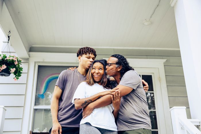 A family of three, with a man and a young man kissing a woman on each cheek, stands on a house porch. A rainbow drawing is visible in the background