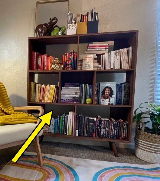 A snug reading corner with a mid-century modern bookshelf stocked with books and decorations, a chair draped with a yellow throw, a plant basket, and a vibrant rug