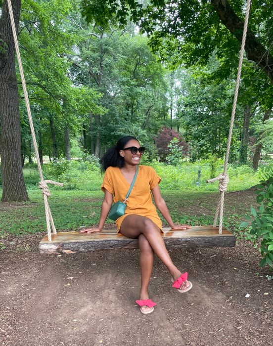 A woman dressed casually sits on a wooden swing in a forested park, smiling while wearing sunglasses and sandals, with a small crossbody bag